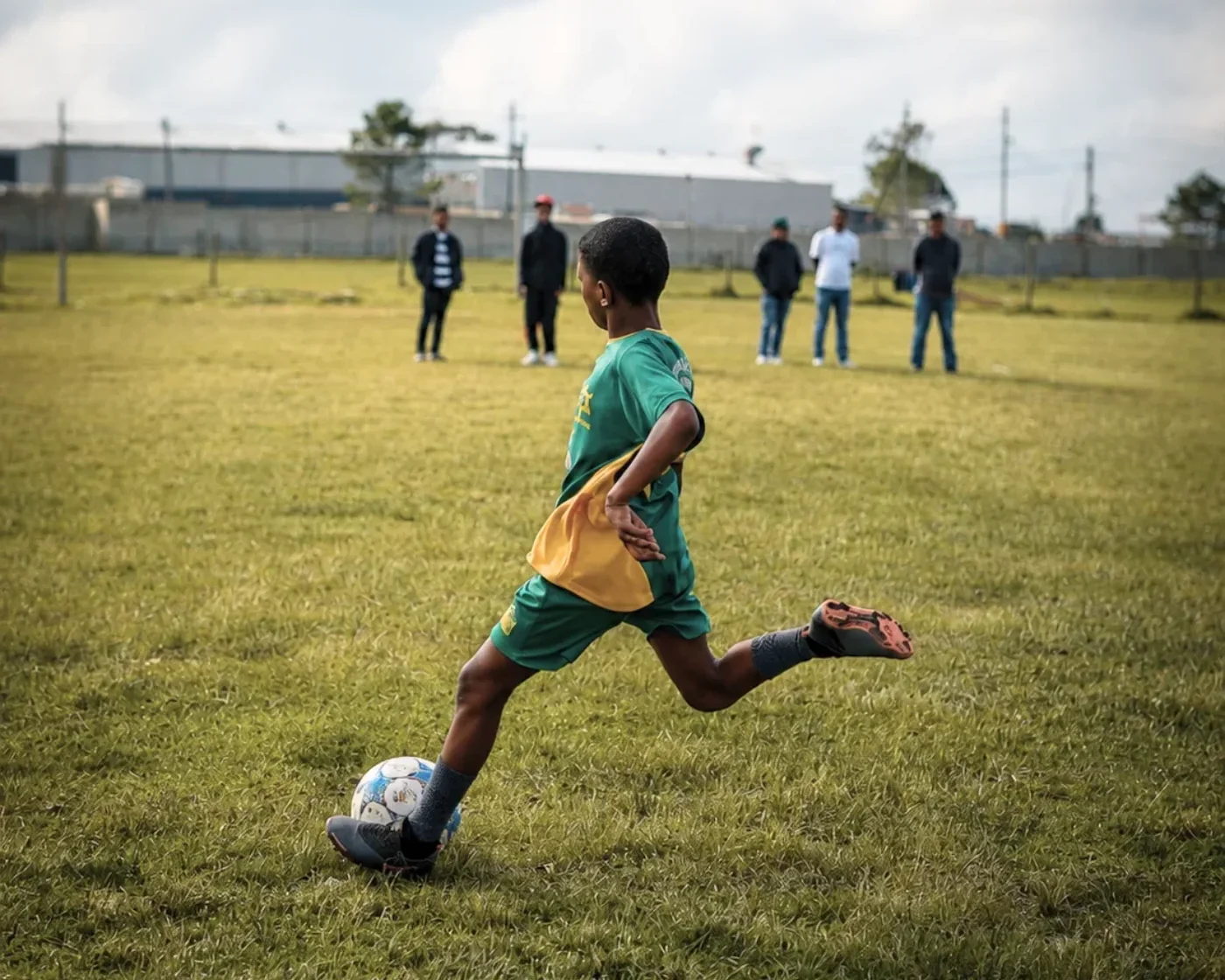 A player in green and gold striking the ball on the grass field, others watching from the touchline